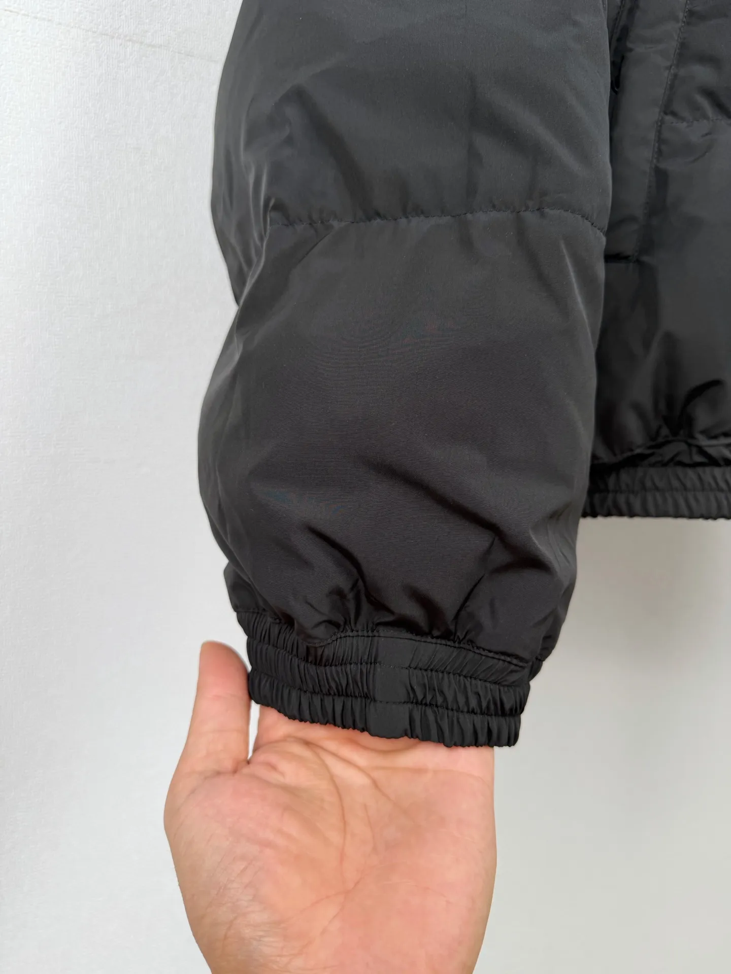 BALENCIAGA / Balenciaga 26SS black and gray reversible ring logo stand-up collar down jacket. This reversible stand-up collar down jacket combines a dual-version appearance with a 90 white duck down lining. One side presents a simple and elegant Black down bread silhouette, and the other side has gray and wine red contrasting panels for a sporty style. Both sides can be worn independently. The craftsmanship is far more complex than ordinary single-sided down garments. In the pattern making and development stage, it is necessary to complete the simultaneous calibration of the two plates. The shoulder slope, armhole arc, zipper placket, and hem elastic point of the front and back sides must be perfectly matched. Once there is a slight deviation in the positioning of the cutting pieces, it will lead to misalignment of the pattern and unbalanced length of the garment when worn on both sides. The down side adopts a horizontal segmented compartment design and is filled with high-quality 90 white duck down. The amount of down filling in each down compartment is accurately weighed and distributed to ensure that the down is evenly distributed and avoid local accumulation or gaps of down material. The fabric is made of high-density anti-drilling down base to minimize the risk of down escaping. The brand logo on the left chest adopts fine thermal transfer printing technology, and the positioning and color number have been reviewed and debugged many times. The body is equipped with a special two-way zipper on both sides, which can be opened and closed smoothly on both sides. The placket adopts a sandwich hidden hemming process, and all sewing lines are stored on the inside of the sandwich. There are no unnecessary exposed stitches on both sides, ensuring a clean and neat appearance. The welt pockets on both sides have an interconnected double-sided structure. The three layers of fabric are aligned simultaneously during sewing. The pocket opening cannot leave stitches on either side that would damage the look and feel. The cuffs and hem adopt hidden elastic hemming technology, and the elastic bands are completely wrapped in the interlayer. There are no traces of elastic bands on the front and back, taking into account windproofness, temperature locking and beautiful lines. The sewing production process is extremely difficult. The sewing operation requires penetrating three layers of outer fabric, 90 fleece liner, and reverse woven fabric at one time. Turners have extremely high requirements for controlling the tightness of stitches. If the stitching is too heavy, it will compress the down bin, damage the down structure, and reduce the warmth retention performance; if the stitching is too loose, it is easy to cause jumpers and down drilling problems. Multiple special-shaped pieces in the contrasting area on the back are spliced ​​with White welts, and the dividing seams must be accurately aligned with the Black side seams. After the garment is sewn, the two facades need to be shaped and ironed separately. One side is baked to give the fluffy and full texture of the down, and the other side is calibrated to make the contrasting color dividing lines straight. The entire production process implements double quality inspection standards, which takes longer, and ultimately allows one piece of clothing to unlock two styles. Size:XS S M L